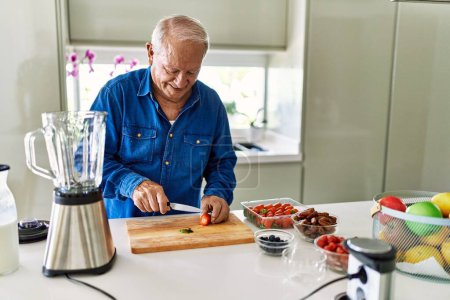 Foto de Hombre mayor sonriendo confiado cortando fresa en la cocina - Imagen libre de derechos