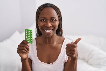 Foto de Mujer afroamericana sosteniendo píldoras anticonceptivas sonriendo feliz y positivo, pulgar hacia arriba haciendo excelente y signo de aprobación - Imagen libre de derechos
