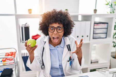 Photo for Black doctor woman with curly hair holding green apple celebrating victory with happy smile and winner expression with raised hands - Royalty Free Image