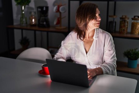 Foto de Mujer hispana de mediana edad usando portátil en casa por la noche mirando al costado, pose de perfil relajado con cara natural con sonrisa confiada. - Imagen libre de derechos
