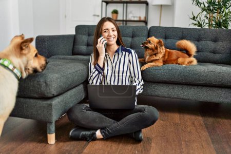 Photo for Young hispanic woman using laptop and talking on the smartphone sitting on floor with dogs at home - Royalty Free Image