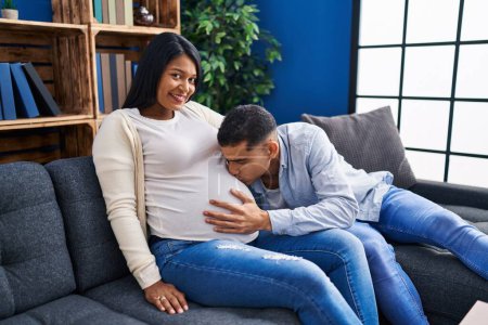 Téléchargez les photos : Jeune couple latin attendant bébé baisers ventre assis sur canapé à la maison - en image libre de droit