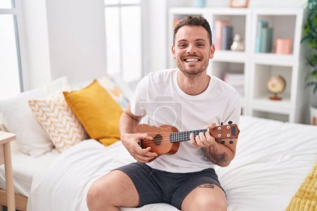 Téléchargez les photos : Jeune homme hispanique jouant ukulele assis sur le lit dans la chambre - en image libre de droit