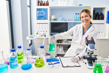 Photo for Young hispanic woman scientist measuring liquid write on document at laboratory - Royalty Free Image