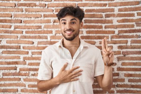 Photo for Arab man with beard standing over bricks wall background smiling swearing with hand on chest and fingers up, making a loyalty promise oath - Royalty Free Image