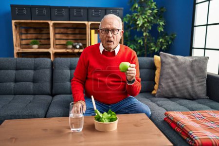 Photo for Senior man with grey hair eating salad and green apple scared and amazed with open mouth for surprise, disbelief face - Royalty Free Image