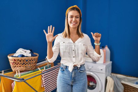 Foto de Joven mujer rubia en la sala de lavandería mostrando y señalando hacia arriba con los dedos número seis mientras sonríe confiado y feliz. - Imagen libre de derechos
