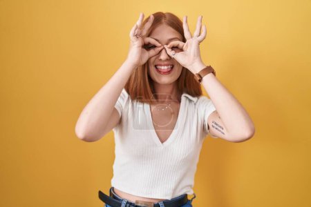Foto de Young redhead woman standing over yellow background doing ok gesture like binoculars sticking tongue out, eyes looking through fingers. crazy expression. - Imagen libre de derechos