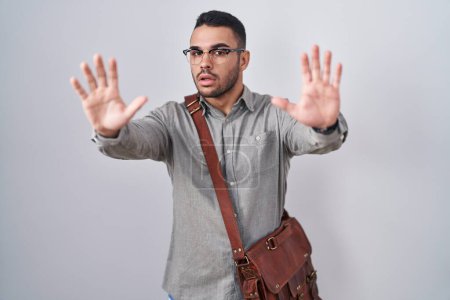 Téléchargez les photos : Jeune homme hispanique portant une valise faisant un geste d'arrêt avec les paumes des mains, expression de colère et de frustration - en image libre de droit