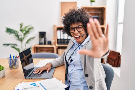 Photo for Black woman with curly hair wearing call center agent headset at the office doing stop gesture with hands palms, angry and frustration expression - Royalty Free Image
