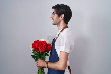 Foto de Young hispanic man holding bouquet of white and red roses looking to side, relax profile pose with natural face and confident smile. - Imagen libre de derechos