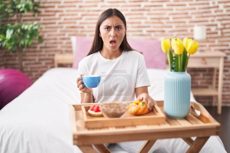 Photo for Young hispanic woman eating pastries for breakfast sitting on the bed in shock face, looking skeptical and sarcastic, surprised with open mouth - Royalty Free Image