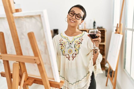 Young hispanic woman drinking wine drawing at art studio