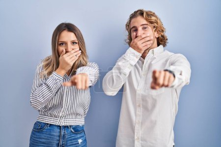Téléchargez les photos : Jeune couple debout sur fond bleu riant de vous, pointant du doigt la caméra avec la main sur la bouche, expression de honte - en image libre de droit