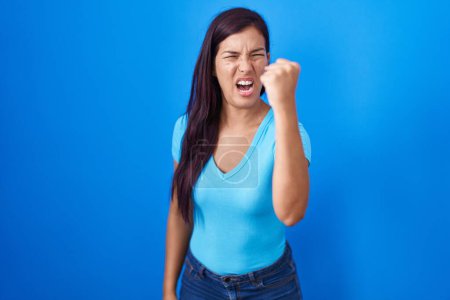 Téléchargez les photos : Jeune femme hispanique debout sur fond bleu en colère et fou levant le poing frustré et furieux tout en criant de colère. rage et concept agressif. - en image libre de droit