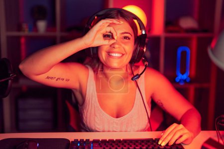 Foto de Mujer rubia joven jugando videojuegos con auriculares haciendo buen gesto con la mano sonriente, los ojos mirando a través de los dedos con la cara feliz. - Imagen libre de derechos