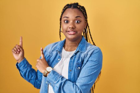 Photo for African american woman with braids standing over yellow background pointing aside worried and nervous with both hands, concerned and surprised expression - Royalty Free Image