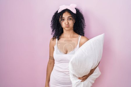 Téléchargez les photos : Hispanic woman with curly hair wearing sleep mask and pajama holding pillow puffing cheeks with funny face. mouth inflated with air, crazy expression. - en image libre de droit