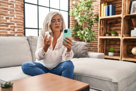 Téléchargez les photos : Femme d'âge moyen utilisant un smartphone tenant des pilules à la maison - en image libre de droit