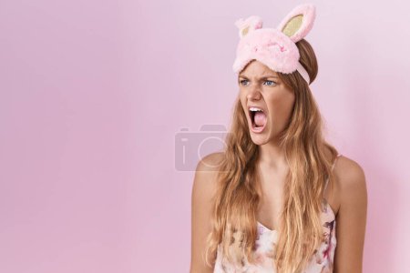 Foto de Mujer caucásica joven con máscara de dormir y pijama enojado y loco gritando frustrado y furioso, gritando con ira. rabia y concepto agresivo. - Imagen libre de derechos