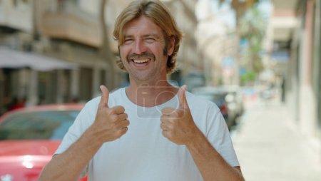 Foto de Young blond man smiling confident doing ok sign with thumbs up at street - Imagen libre de derechos