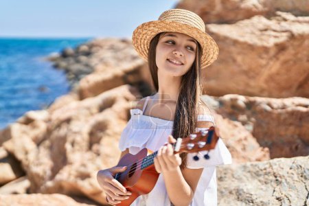 Téléchargez les photos : Adorable fille touristique souriant confiant jouer ukulele au bord de la mer - en image libre de droit