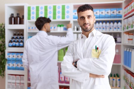 Téléchargez les photos : Two hispanic men pharmacists smiling confident standing with arms crossed gesture at pharmacy - en image libre de droit