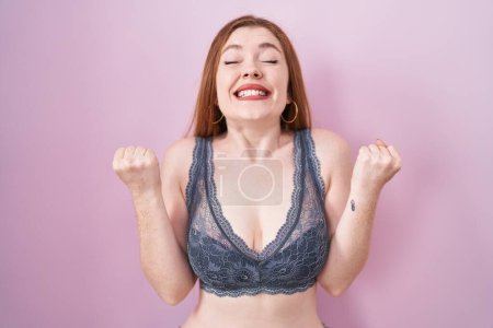 Téléchargez les photos : Redhead woman wearing lingerie over pink background excited for success with arms raised and eyes closed celebrating victory smiling. winner concept. - en image libre de droit