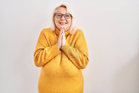 Foto de Mujer caucásica de mediana edad con gafas de pie sobre el fondo rezando con las manos juntas pidiendo perdón sonriendo confiado. - Imagen libre de derechos