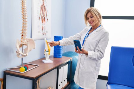 Photo for Middle age blonde woman wearing physiotherapist uniform using smartphone pointing to anatomical model of knee at physiotherapy clinic - Royalty Free Image