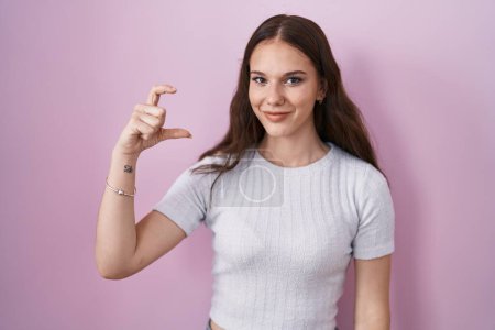 Photo for Young hispanic girl standing over pink background smiling and confident gesturing with hand doing small size sign with fingers looking and the camera. measure concept. - Royalty Free Image