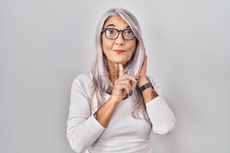 Foto de Mujer de mediana edad con el pelo gris de pie sobre fondo blanco pensando concentrado en la duda con el dedo en la barbilla y mirando hacia arriba preguntándose - Imagen libre de derechos