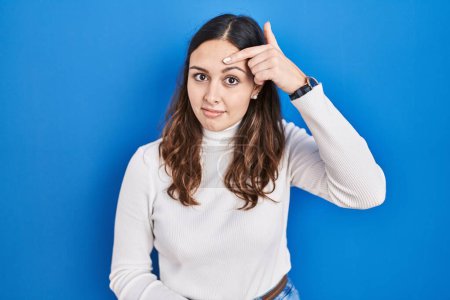 Photo for Young hispanic woman standing over blue background pointing unhappy to pimple on forehead, ugly infection of blackhead. acne and skin problem - Royalty Free Image