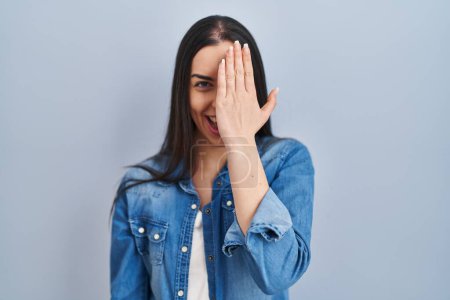 Téléchargez les photos : Femme hispanique debout sur fond bleu couvrant un oeil avec la main, sourire confiant sur le visage et l'émotion surprise. - en image libre de droit