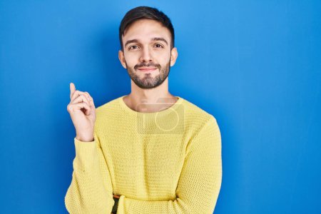 Photo for Hispanic man standing over blue background pointing with hand finger to the side showing advertisement, serious and calm face - Royalty Free Image