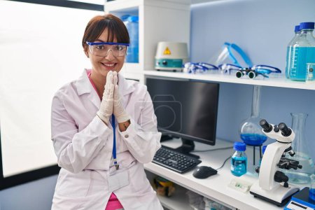 Photo for Young brunette woman working at scientist laboratory praying with hands together asking for forgiveness smiling confident. - Royalty Free Image