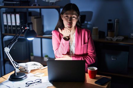 Téléchargez les photos : Chinoise jeune femme travaillant au bureau la nuit en regardant la caméra souffler un baiser avec la main sur l'air étant belle et sexy. expression d'amour. - en image libre de droit