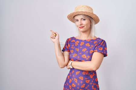 Téléchargez les photos : Jeune femme caucasienne portant des fleurs robe et chapeau d'été pointant avec le doigt de la main sur le côté montrant la publicité, visage sérieux et calme - en image libre de droit
