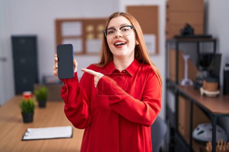 Téléchargez les photos : Redhead woman working at the office showing smartphone screen smiling happy pointing with hand and finger - en image libre de droit