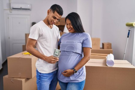 Photo for Young latin couple expecting baby holding dollars standing at new home - Royalty Free Image