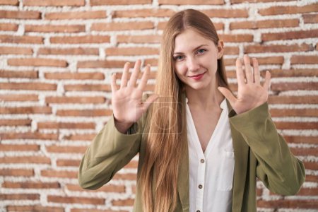 Téléchargez les photos : Jeune femme caucasienne debout sur le fond de mur de briques montrant et pointant vers le haut avec les doigts numéro dix tout en souriant confiant et heureux. - en image libre de droit