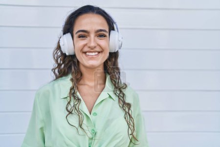 Foto de Mujer hispana joven escuchando música usando auriculares que se ven positivos y felices de pie y sonriendo con una sonrisa confiada mostrando los dientes - Imagen libre de derechos
