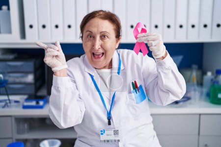 Photo for Senior woman with glasses working at scientist laboratory holding pink ribbon smiling happy pointing with hand and finger to the side - Royalty Free Image