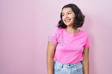 Foto de Mujer hispana joven de pie sobre fondo rosa mirando hacia otro lado con sonrisa en la cara, expresión natural. riendo confiado. - Imagen libre de derechos