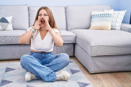 Téléchargez les photos : Young caucasian woman sitting on the floor at the living room shouting angry out loud with hands over mouth - en image libre de droit