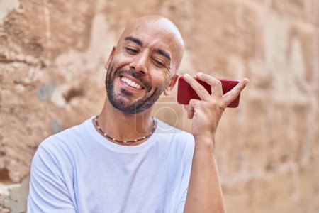 Téléchargez les photos : Jeune homme chauve souriant confiant écoute message audio par le smartphone sur fond de pierre isolée - en image libre de droit