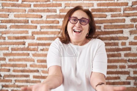 Téléchargez les photos : Femme âgée avec des lunettes debout sur le mur de briques souriant joyeuse offrant des mains donnant assistance et acceptation. - en image libre de droit