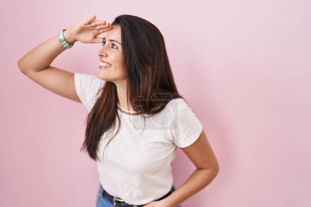 Photo for Young brunette woman standing over pink background very happy and smiling looking far away with hand over head. searching concept. - Royalty Free Image