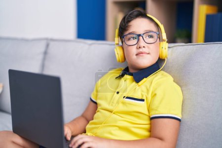 Téléchargez les photos : Adorable garçon hispanique utilisant ordinateur portable et écouteurs assis sur le canapé à la maison - en image libre de droit