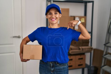 Foto de Middle age brunette woman working wearing delivery uniform and cap looking confident with smile on face, pointing oneself with fingers proud and happy. - Imagen libre de derechos